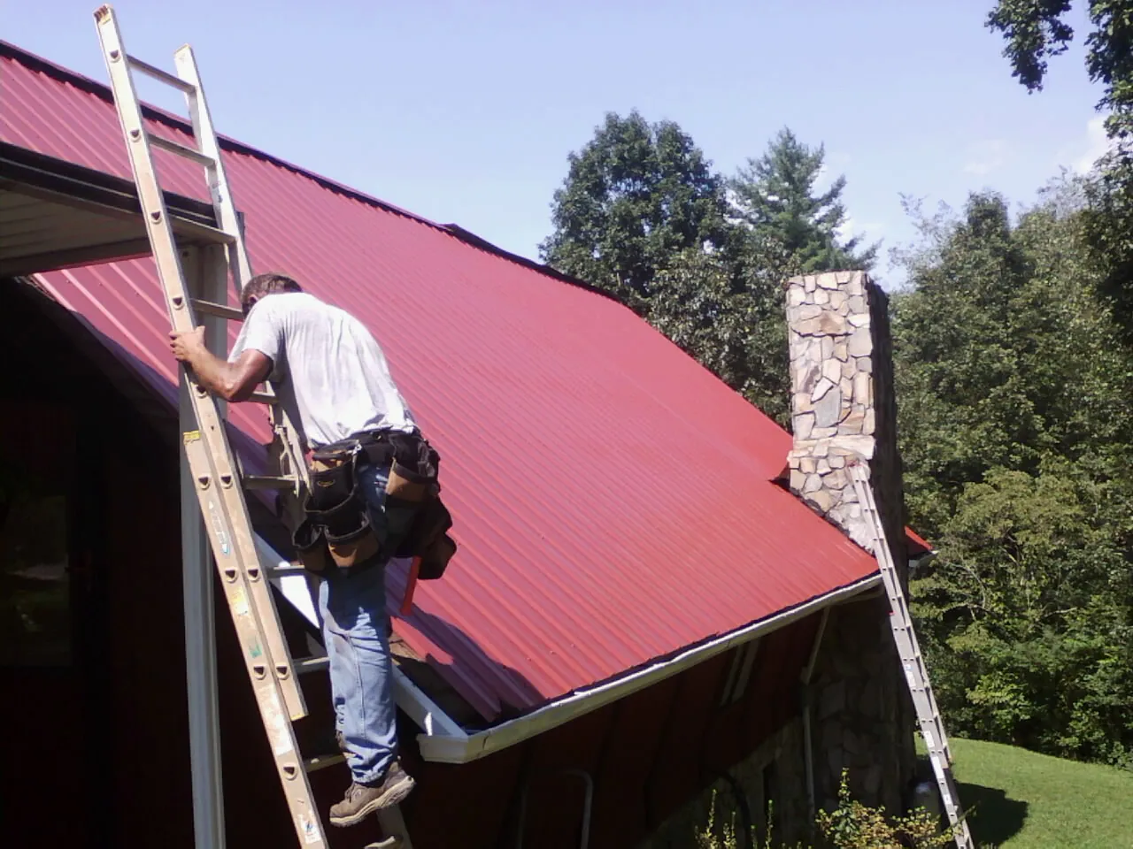 Roofer on a ladder beside a red metal roof and stone chimney — Soffit & Fascia Repair by Roofing Brothers in Stockertown